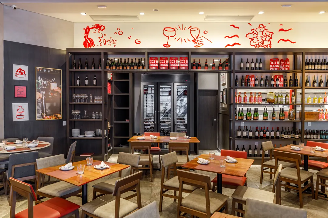 Salão da ICI Brasserie com adega em estante metálica e ilustrações vermelhas na parede