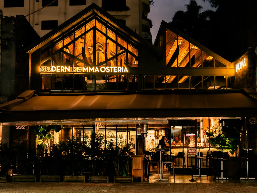 Fachada do MoMa (Modern Mamma Osteria), em estrutura de madeira e vidro, à noite