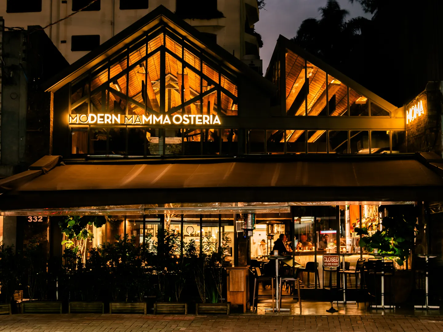 Fachada do MoMa (Modern Mamma Osteria), em estrutura de madeira e vidro, à noite