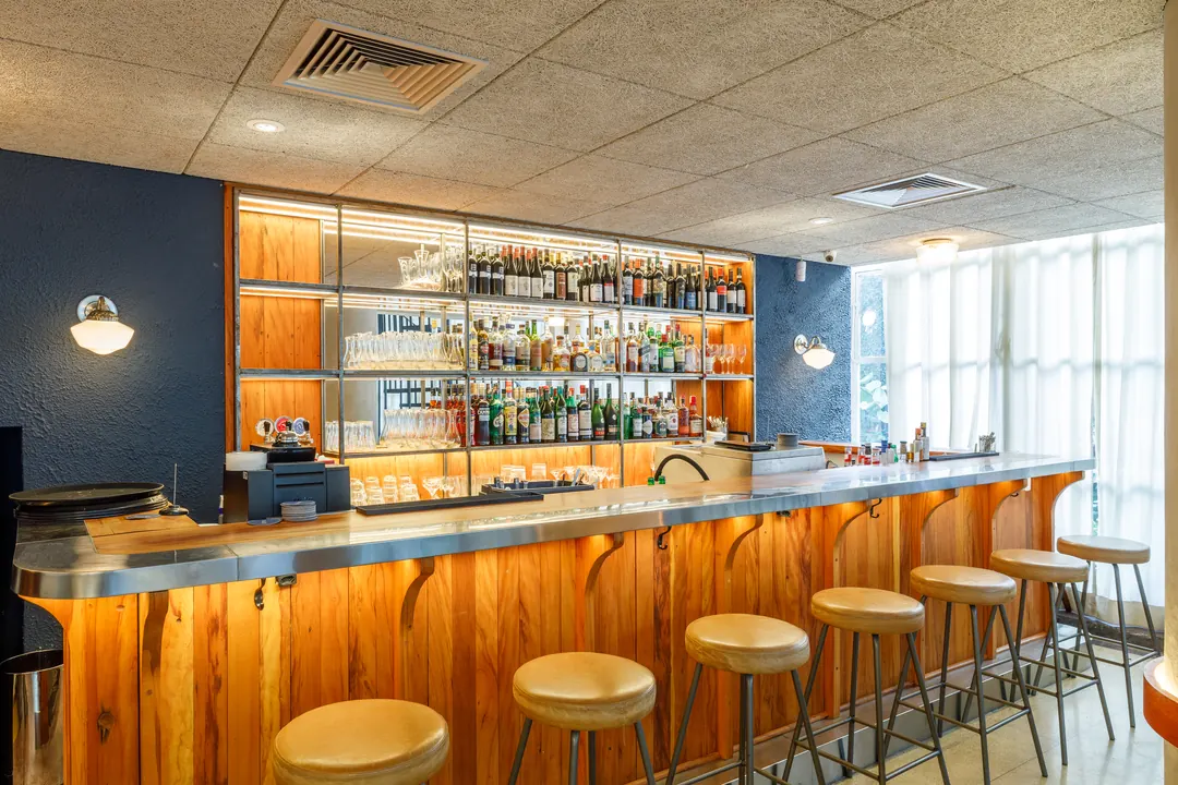 Balcão do bar em madeira com bancos altos e prateleiras de garrafas